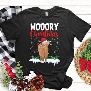 May include: Black t-shirt with the text "MOOORY Christmas" in white and red. A brown Highland cow wearing a Santa hat and Christmas lights is in the center. Snowflakes and a snow-covered ground complete the festive design.