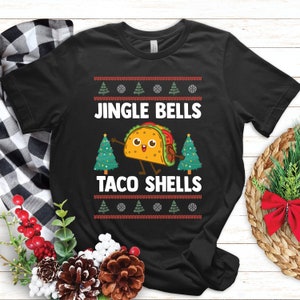 May include: Black t-shirt featuring the text "JINGLE BELLS TACO SHELLS" and a cartoon taco. The design includes Christmas trees and a festive sweater-style border. The shirt is made of a soft material.