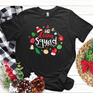 Może przedstawiać: Czarna koszulka z świątecznym wzorem. Koszulka zawiera napis "HR Christmas Squad" w fantazyjnej czcionce, otoczony świątecznymi ilustracjami, takimi jak ozdoby, czapka Świętego Mikołaja i piernikowy ludzik. Idealny prezent świąteczny.