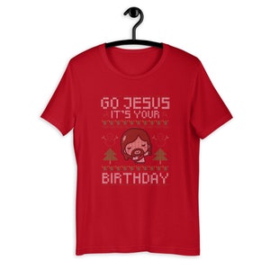 May include: Red t-shirt with the text "GO JESUS IT'S YOUR BIRTHDAY" in a pixelated font. The design includes a cartoon Jesus with a beard, Christmas trees, and snowmen. The shirt is hanging on a black hanger.