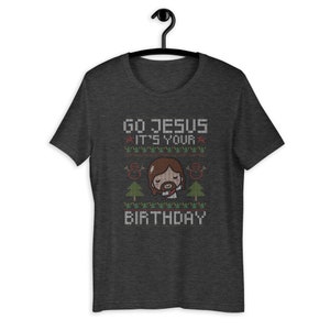May include: Dark gray t-shirt with a graphic design. The design features the text "GO JESUS IT'S YOUR BIRTHDAY" in a pixelated font, along with an illustration of Jesus and Christmas trees. The shirt is hanging on a black hanger.