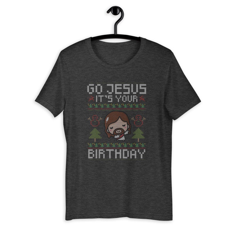 May include: Dark gray t-shirt with a graphic design. The design features the text "GO JESUS IT'S YOUR BIRTHDAY" in a pixelated font, along with an illustration of Jesus and Christmas trees. The shirt is hanging on a black hanger.