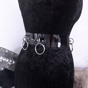 May include: A black, glossy belt with silver metal rings and studs. The belt is displayed on a black velvet mannequin. The belt has three large silver rings and several smaller silver studs.