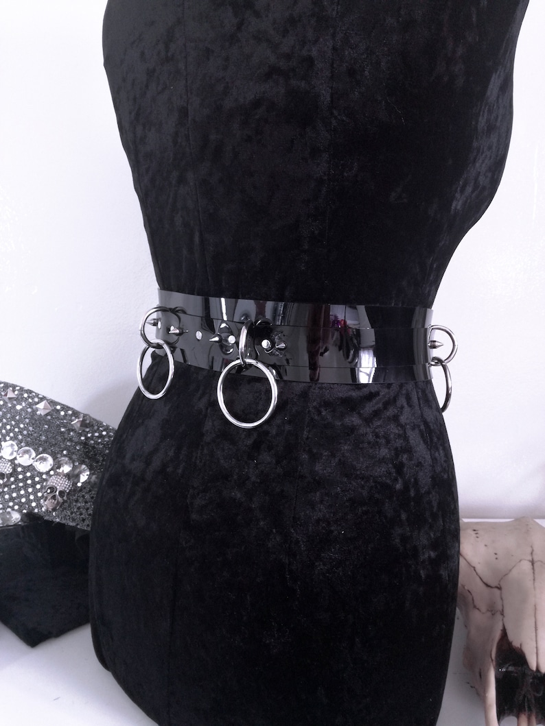 May include: A black, glossy belt with silver metal rings and studs. The belt is displayed on a black velvet mannequin. The belt has three large silver rings and several smaller silver studs.