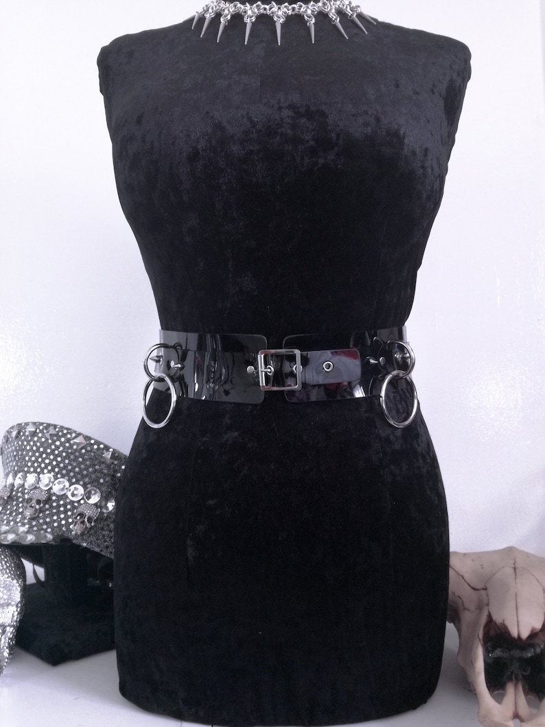 May include: A black velvet mannequin displays a shiny black belt with silver rings and a buckle. A silver spiked choker necklace is at the neck. A skull and a hat with skull embellishments are in the background.