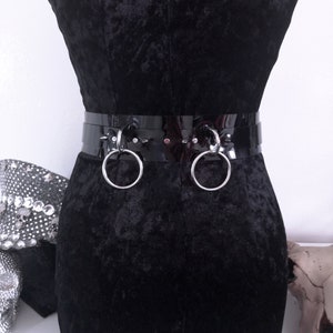May include: A black patent leather belt with two large silver metal rings. The belt is worn on a black velvet form. The belt has a shiny, reflective surface and is a bold fashion accessory.