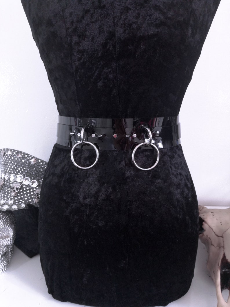 May include: A black patent leather belt with two large silver metal rings. The belt is worn on a black velvet form. The belt has a shiny, reflective surface and is a bold fashion accessory.
