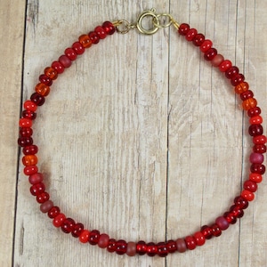 May include: A beaded bracelet featuring a variety of red and orange beads. The beads are strung together with a gold-colored clasp. The bracelet is displayed on a light brown wooden surface.