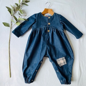 Peut inclure: Une combinaison bébé en denim bleu avec des manches longues et une fermeture à boutons. La combinaison a un patch en tissu à carreaux sur la jambe droite et une jupe plissée.