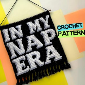 Può includere: Un arazzo da parete all'uncinetto nero con le parole "IN MY NAP ERA" in bianco, con lettere in stile 3D. Il bordo inferiore ha una frangia. Le parole "CROCHET PATTERN" sono in un rettangolo verde e blu.