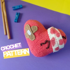 Puede incluir: Un corazón rojo de ganchillo hecho a mano con una venda y un parche. El corazón tiene una cara cosida con una expresión triste. Las palabras "CROCHET PATTERN" están en una etiqueta amarilla y blanca.