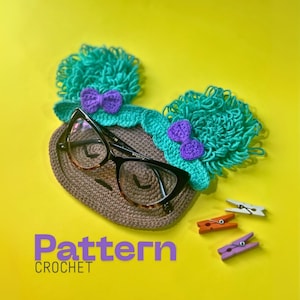 Könnte beinhalten: Ein gehäkelter Brillenhalter mit einem skurrilen Design, der türkisfarbene Haarbüschel, lila Schleifen und eine Brille zeigt. Der Halter ist braun, und das Bild enthält den Text "Pattern CROCHET".