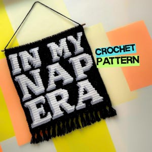以下が含まれることがあります： ブロック体で「IN MY NAP ERA」という文字が編み込まれた、黒と白のクロシェ編みの壁掛け。黒い紐で木製の棒から吊るされており、下端には黒いフリンジが付いています。画像には「CROCHET PATTERN」というテキストも含まれています。