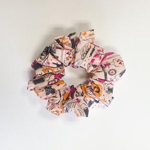 Puede incluir: Una goma para el pelo rosa claro con un estampado de Halloween. La tela presenta ilustraciones de calabazas, arañas y otros elementos espeluznantes en negro, naranja y rosa. La goma es fruncida y circular.