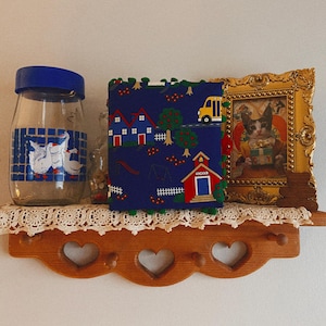 May include: A wooden shelf with a decorative lace doily, holding a glass jar with a blue lid and goose design, a blue fabric item with a schoolhouse pattern, and a framed picture of a cat.