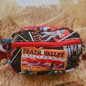 May include: A quilted fanny pack with a red zipper and strap. The bag features a colorful collage of images and the text "DEATH VALLEY CALIFORNIA" on the front. The background is a cream-colored lace.