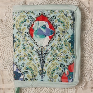 May include: A quilted book cover with a light blue background featuring a repeating floral pattern with dog portraits in red frames. The cover is edged with a light blue fabric and has a green ribbon closure.