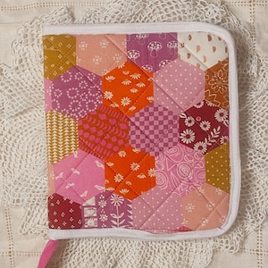 May include: A handmade, quilted baby bonnet featuring a patchwork design of hexagonal fabric pieces in shades of pink, orange, and gold. The bonnet has a white trim and a pink tie. The fabric pieces feature floral and geometric patterns.