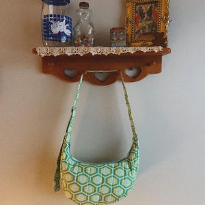 Könnte beinhalten: Eine kleine, gesteppte Umhängetasche mit einem geometrischen Muster aus grünen, gelben und blauen Sechsecken. Die Tasche hängt an einem Holzregal mit herzförmigen Ausschnitten. Auf dem Regal befinden sich auch ein Glas, eine bärenförmige Flasche und ein gerahmtes Bild.