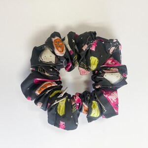 Puede incluir: Una scrunchie negra con un patrón repetido de ilustraciones coloridas. El patrón incluye imágenes de botellas, vasos y otros artículos de bar. La scrunchie está hecha de una tela suave y está diseñada para sujetar el cabello en su lugar.