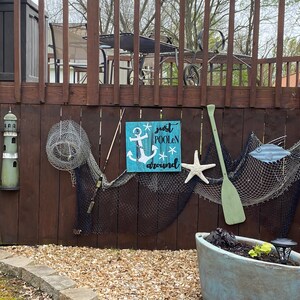 Puede incluir: Un letrero de madera con fondo azul y letras blancas que dice "just poolin' around". El letrero presenta un ancla blanca y estrellas. El letrero est&aacute; colgado en una valla de madera con otras decoraciones n&aacute;uticas, incluyendo un faro, redes de pesca y una pala de madera.