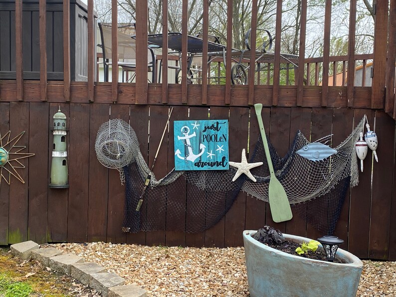 Puede incluir: Un letrero de madera con fondo azul y letras blancas que dice "just poolin' around". El letrero presenta un ancla blanca y estrellas. El letrero est&aacute; colgado en una valla de madera con otras decoraciones n&aacute;uticas, incluyendo un faro, redes de pesca y una pala de madera.