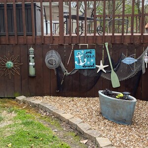 Puede incluir: Un letrero de madera con fondo azul y texto blanco que dice "Just poolin' around". El letrero presenta un ancla blanca y una estrella de mar blanca. El letrero est&aacute; colgado en una valla de madera con una red de pesca, un remo de madera y otras decoraciones n&aacute;uticas.