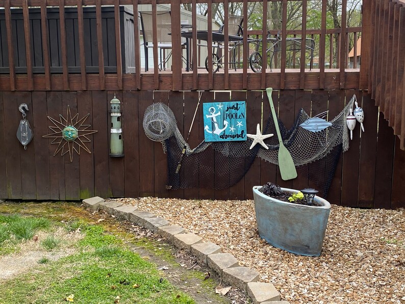 Puede incluir: Un letrero de madera con fondo azul y texto blanco que dice "Just poolin' around". El letrero presenta un ancla blanca y una estrella de mar blanca. El letrero est&aacute; colgado en una valla de madera con una red de pesca, un remo de madera y otras decoraciones n&aacute;uticas.