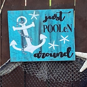 Puede incluir: Un letrero de madera azul con un ancla blanca y el texto "just poolen around" en negro. El letrero tiene un aspecto desgastado y est&aacute; decorado con estrellas de mar blancas.