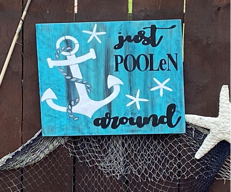 Puede incluir: Un letrero de madera azul con un ancla blanca y el texto "just poolen around" en negro. El letrero tiene un aspecto desgastado y est&aacute; decorado con estrellas de mar blancas.