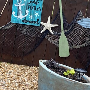 Puede incluir: Un letrero de madera azul con un ancla blanca y el texto "just poolen around". El letrero est&aacute; colgado en una valla de madera marr&oacute;n con una estrella de mar blanca y una pala verde.