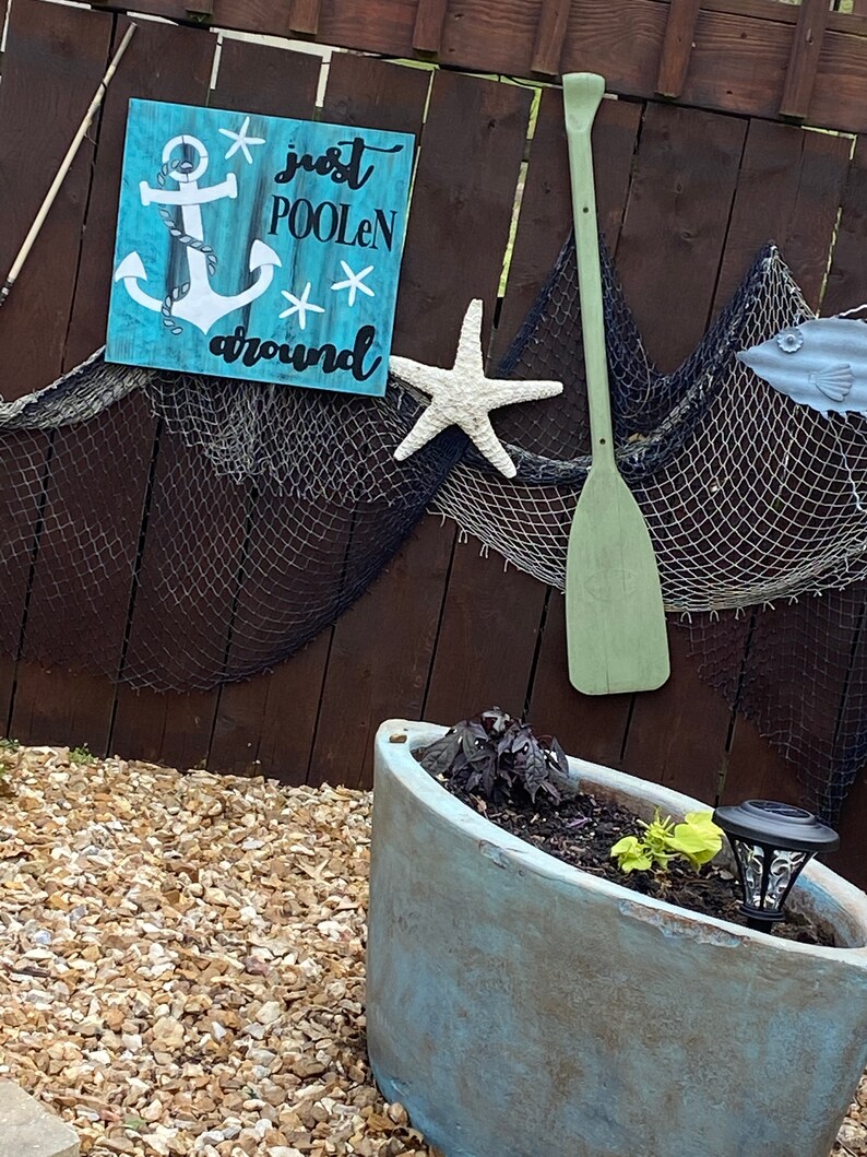Puede incluir: Un letrero de madera azul con un ancla blanca y el texto "just poolen around". El letrero est&aacute; colgado en una valla de madera marr&oacute;n con una estrella de mar blanca y una pala verde.