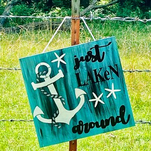 Puede incluir: Un letrero de madera azul turquesa con un ancla blanca y el texto "just laken around" con dos estrellas de mar blancas.
