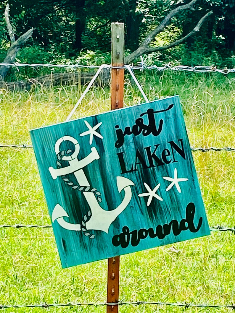 Puede incluir: Un letrero de madera azul turquesa con un ancla blanca y el texto "just laken around" con dos estrellas de mar blancas.