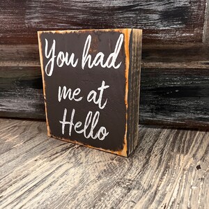 May include: A rustic wooden sign with the text "You had me at Hello" painted in white on a black background.