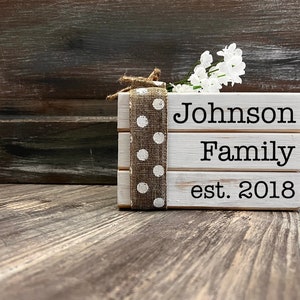 May include: White wooden block sign with the text "Johnson Family est. 2018". The sign is decorated with a brown burlap ribbon with white polka dots and a sprig of white flowers.