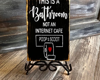 Peut inclure: Une pancarte en bois noir avec du texte blanc qui dit "Ceci est une salle de bain, pas un cybercafé, Poop & Scoot". Un rouleau de papier toilette blanc avec un cœur rouge est dessiné sous le texte. La pancarte est sur un support en métal noir.