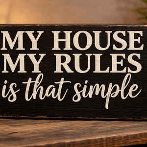 May include: A rectangular black wooden sign with white lettering that reads "MY HOUSE MY RULES is that simple." The sign has a distressed look and sits on a wooden surface. A decorative lantern is in the background.