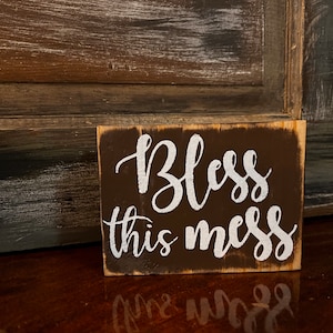 May include: A small wooden sign with a brown background and white lettering that reads "Bless this mess".