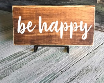 May include: A rectangular wooden sign with the words "be happy" in white script. The sign has a dark brown stain and is displayed on a black metal stand. The background is a light gray wooden surface.