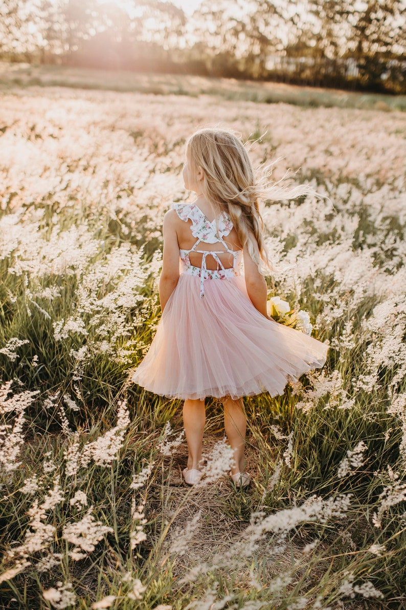 Puede incluir: Una ni&ntilde;a peque&ntilde;a con un vestido de tul rosa con un corpi&ntilde;o de estampado floral se encuentra de pie en un campo de hierba alta. Su cabello es largo y rubio, y lleva un ramo de flores.