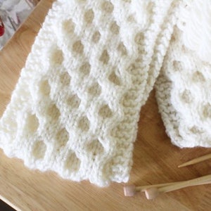 May include: A cream-coloured, open-weave knitted scarf with a honeycomb pattern. The scarf is partially folded on a wooden surface, with a ball of yarn and knitting needles nearby. The scarf is made of a soft, textured yarn.