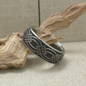 Op de afbeelding: Een zilveren ring met een Keltisch knoopontwerp. De ring ligt op een stuk drijfhout.
