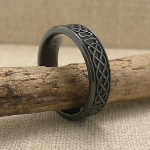 Op de afbeelding: Een donkergrijze ring met een Keltisch knoopmotief. De metalen ring heeft een gepolijste afwerking en het Keltische knoopontwerp is rond de band gegraveerd. De ring wordt gepresenteerd op een stuk hout.