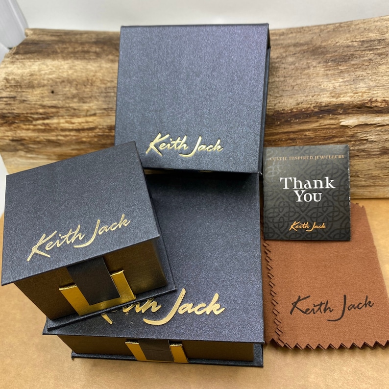 Peut inclure: Plusieurs bo&icirc;tes &agrave; bijoux noires avec l'inscription dor&eacute;e "Keith Jack" sont empil&eacute;es. Une petite carte avec les mots "Thank You" et un motif celtique est visible, ainsi qu'un chiffon de polissage marron.