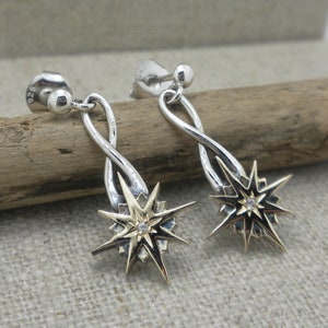 May include: A pair of silver and gold earrings with a starburst design and a small clear stone in the center. The earrings are hanging from a twisted silver wire.