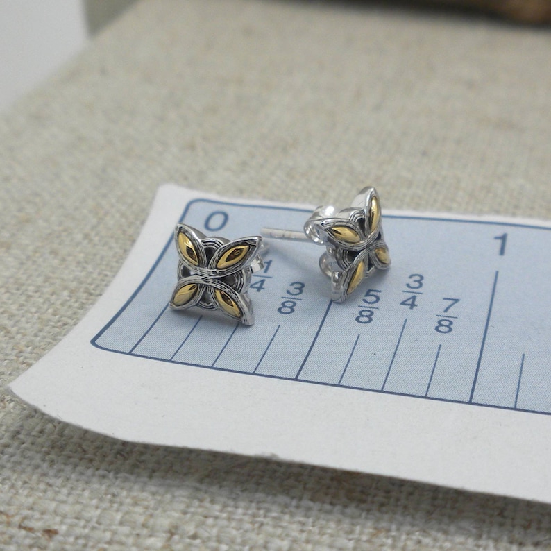 Peut inclure: Une paire de boucles d'oreilles &agrave; clous en argent avec un motif floral dor&eacute;. Les boucles d'oreilles sont de forme carr&eacute;e avec quatre p&eacute;tales dor&eacute;s. Les boucles d'oreilles sont sur une r&egrave;gle blanche et bleue, avec des mesures en centim&egrave;tres.