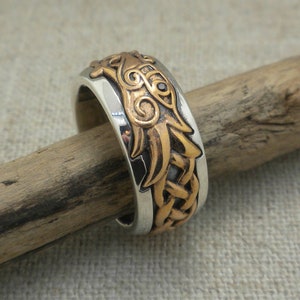 Puede incluir: Un anillo de plata y cobre con un diseño de dragón. El dragón está en el centro del anillo y está hecho de cobre. El resto del anillo es de plata.