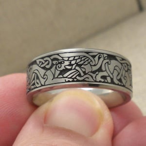 Op de afbeelding: Een zilverkleurige ring met een gedetailleerd, gegraveerd ontwerp met verweven dierfiguren en Keltische knoopmotieven. De ring heeft een brede band en een gepolijste afwerking. Het ontwerp is donker tegen de zilveren achtergrond.