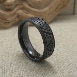 Op de afbeelding: Een zwarte ring met een Keltisch knoopmotief gegraveerd rond de band. De ring heeft een gepolijste afwerking en het woord "GEIF" is aan de binnenkant gegraveerd. Gepresenteerd op een neutrale achtergrond.